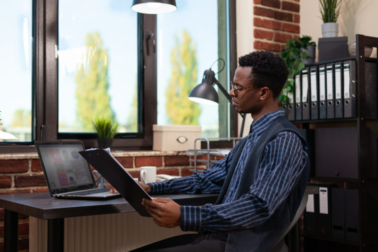 Entrepreneur looking at laptop screen with sales results holding clipboard with bussiness charts